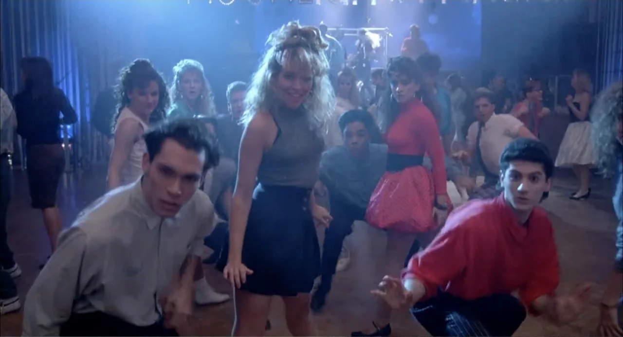 Teen Witch (1989) movie still: medium — A crowded high school gymnasium at a dance with blue stage lights in back. A small group of teenagers dances…