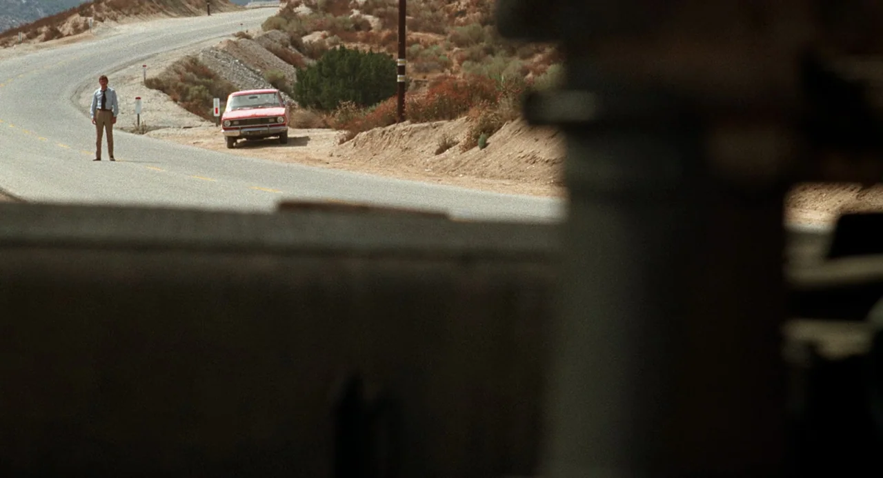 Duel (1971) movie still: wide — A winding asphalt road cuts through a dry, hilly desert landscape with sparse vegetation. A man in a dress shirt and…