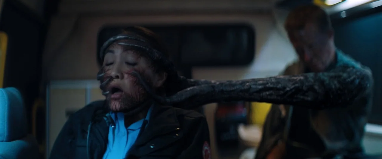 Venom (2018) movie still: medium — A dimly lit, cramped medical vehicle interior with blue-tinted shadows and low visibility. An alien-infected male…