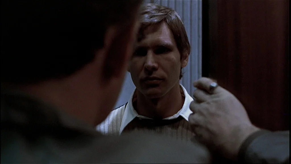 The Conversation (1974) movie still: medium — A dark bathroom interior with a reflective surface visible in the center frame. A man looks directly…