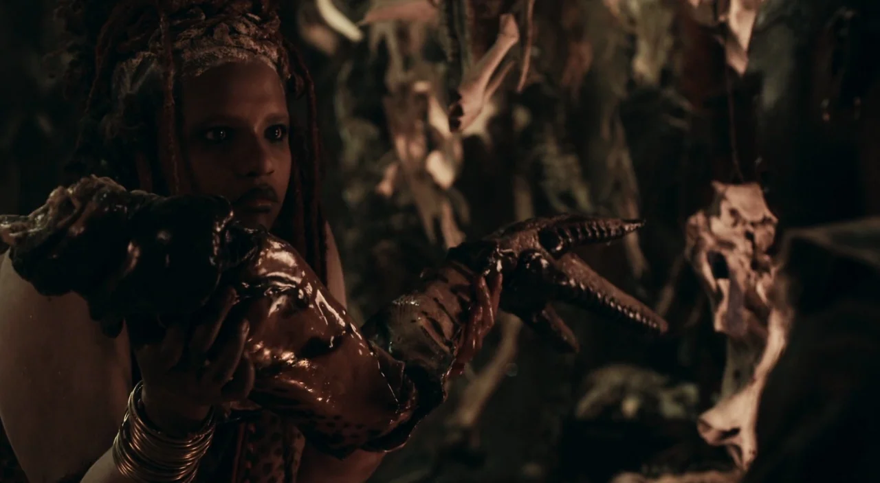 District 9 (2009) movie still: medium — A dark, cramped room contains numerous animal bones hanging from the low ceiling. A man holds a monstrous,…