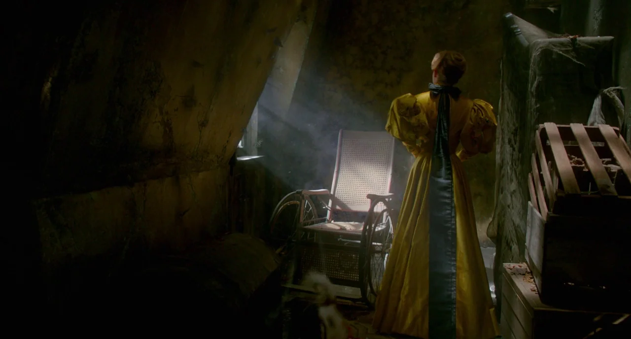 Crimson Peak (2015) movie still: fullbody — A dimly lit, dusty attic room is cluttered with old wooden crates and cobwebs. A woman in a yellow…
