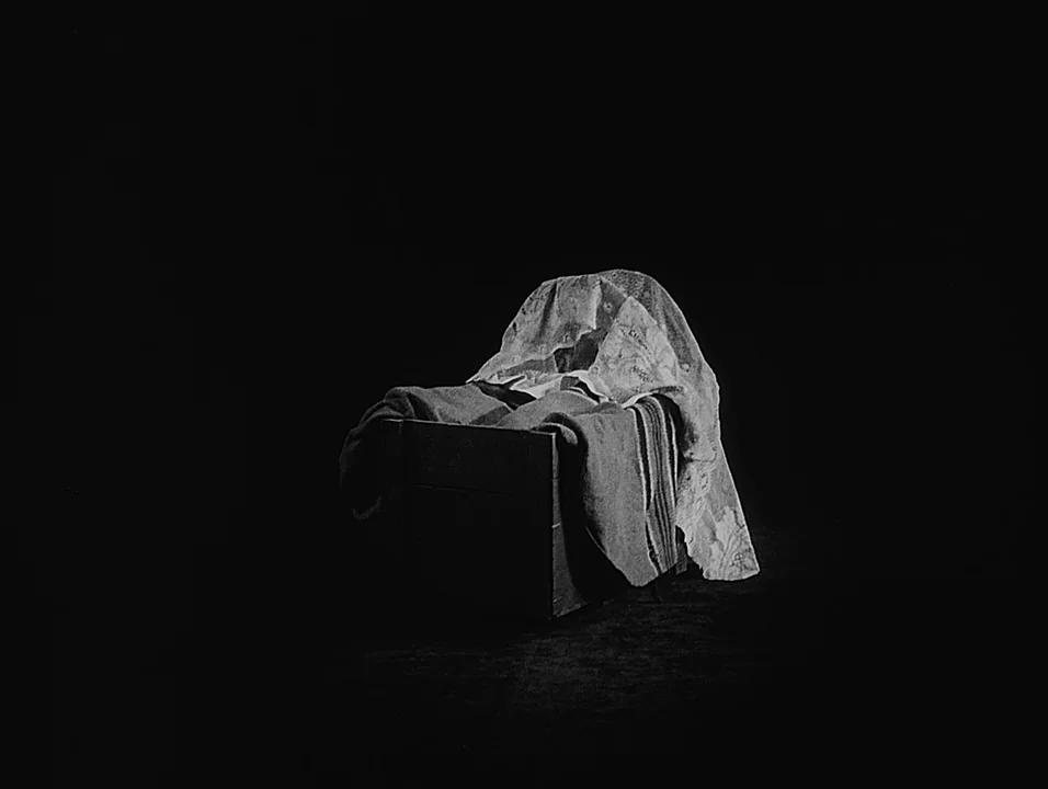 Coeur Fidele (1923) movie still: medium — A simple wooden cradle sits on a dark floor against an entirely black background. A white patterned lace…