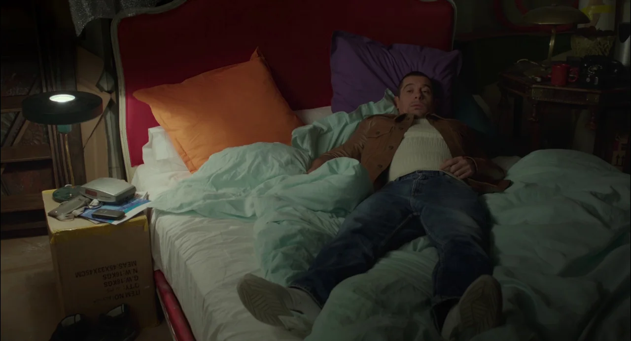 Abuse of Weakness (2013) movie still: medium — A man lies on a bed with mismatched pillows in a dimly lit space. He rests on his back with hands…