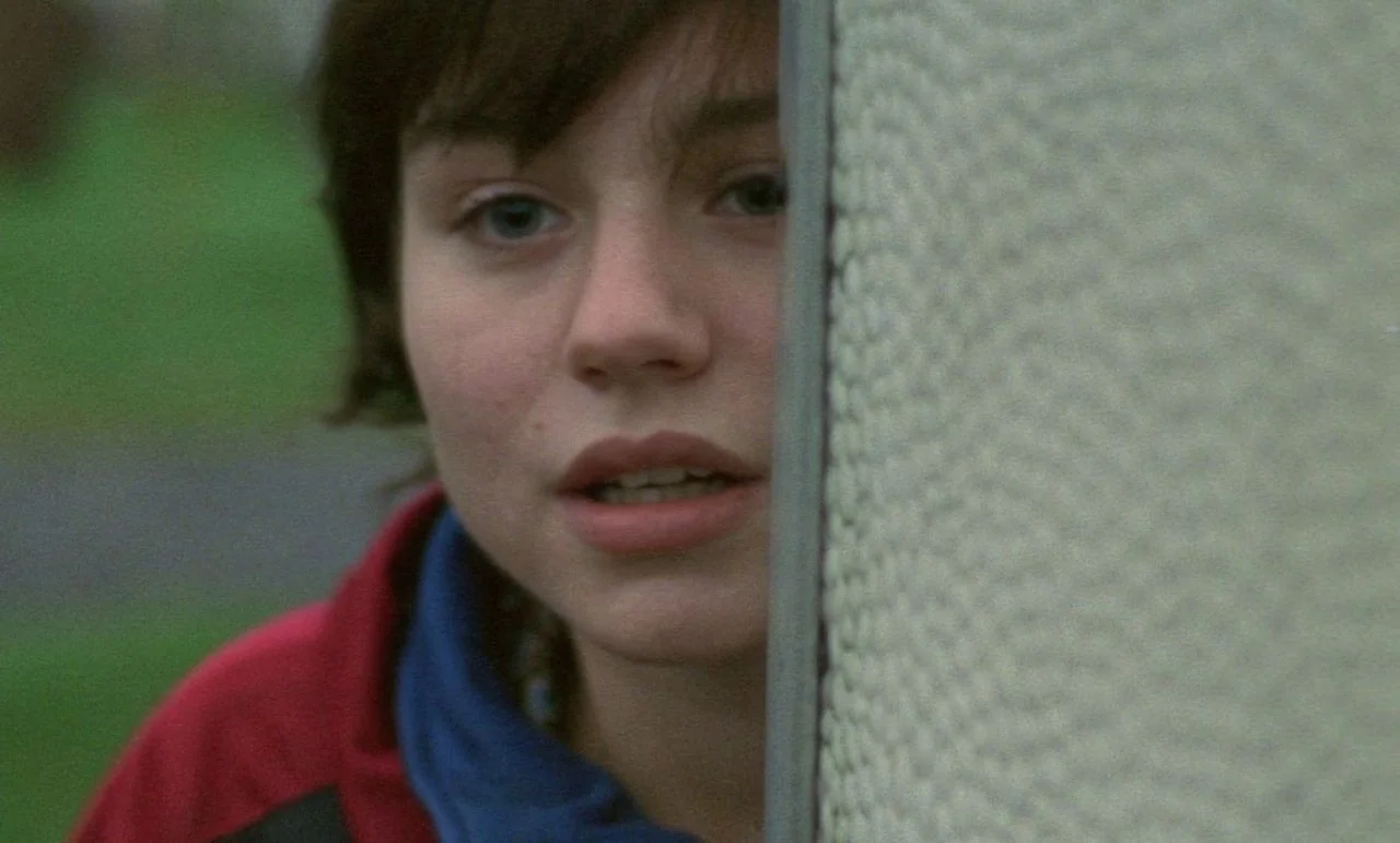 Rosetta (1999) movie still: closeup — A teenage girl looks out from behind the textured gray concrete corner of a building. Green foliage is visible…