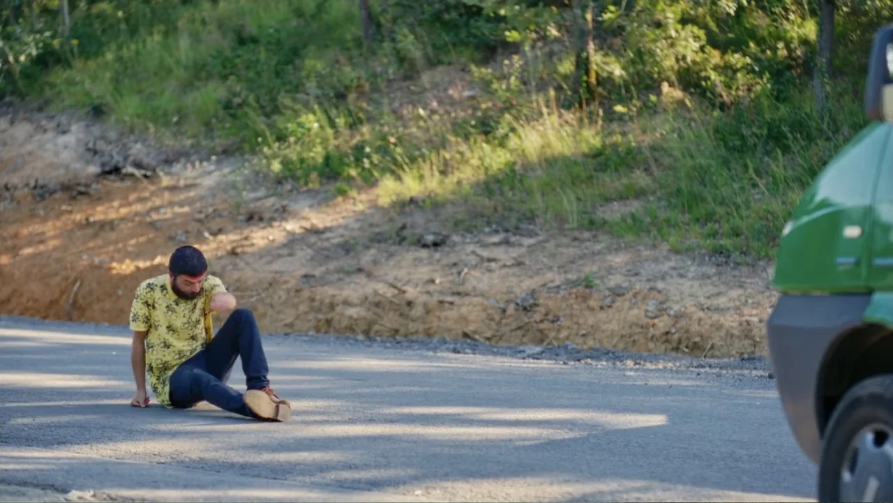 Paranormal Cuma (2024) movie still: medium — A man sits on the side of a rural paved road near a green vehicle. The man has a bandaged forehead and…