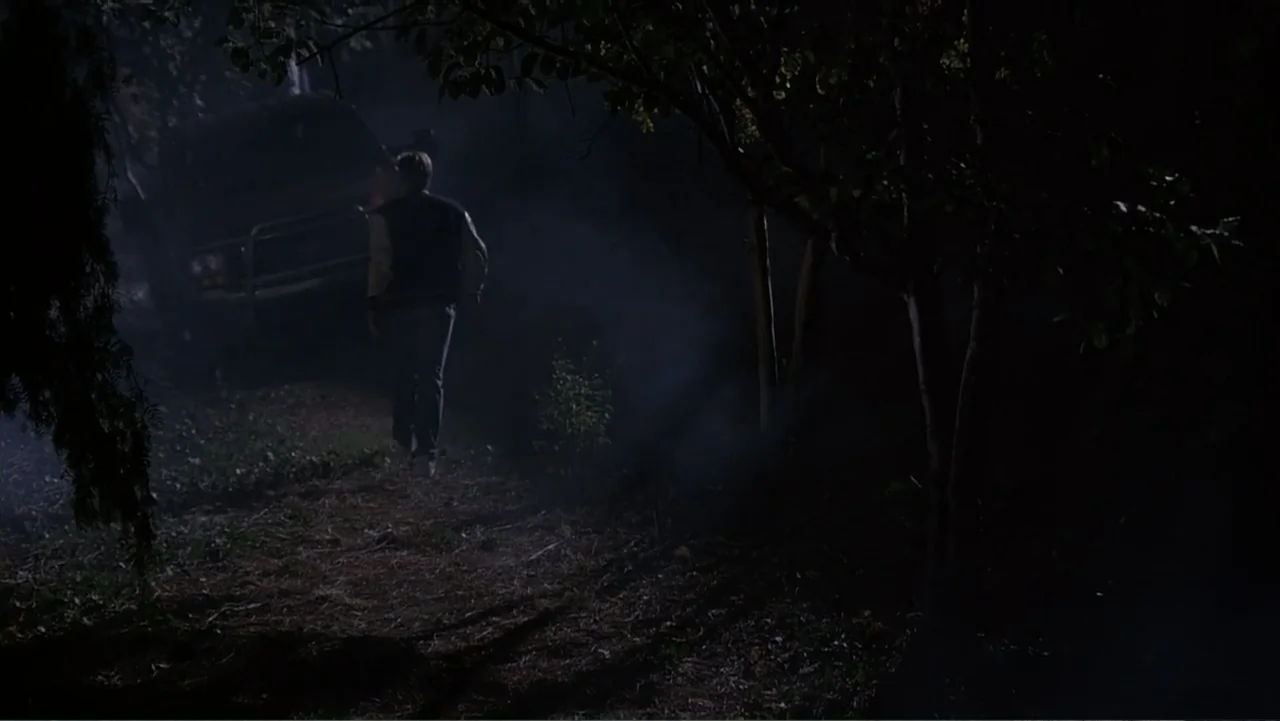 Crisis Point (2012) movie still: wide — A dark wooded path at night with a large vehicle partially visible behind trees. A lone male figure walks…