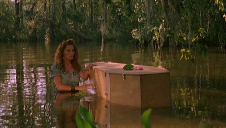 Highway 61 (1991) movie still: medium — A woman stands waist-deep in murky swamp water near a wooden coffin. She rests her hands on the lid of the…