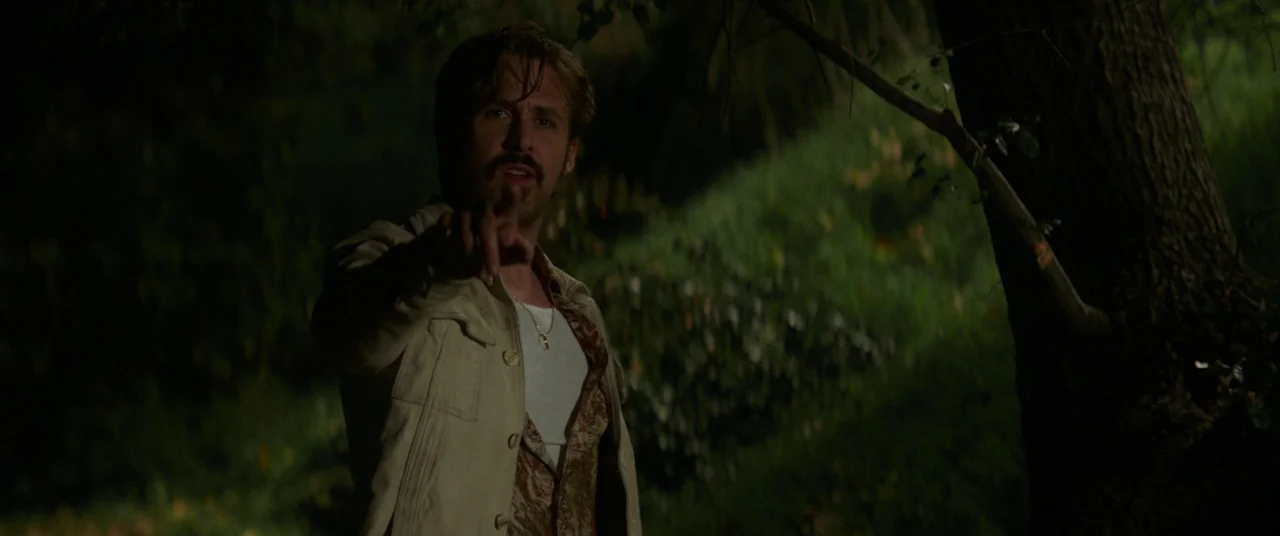 The Nice Guys (2016) movie still: medium — A dark outdoor space at night with trees and overgrown grassy hills in background. A man stands in the…