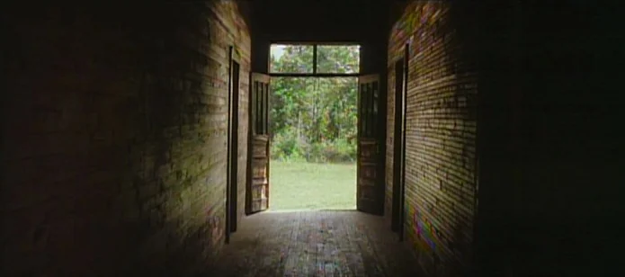 The Neon Bible (1995) movie still: establishing — A dark, narrow interior hallway made of horizontal wooden planks leads to an exit. An open doorway…