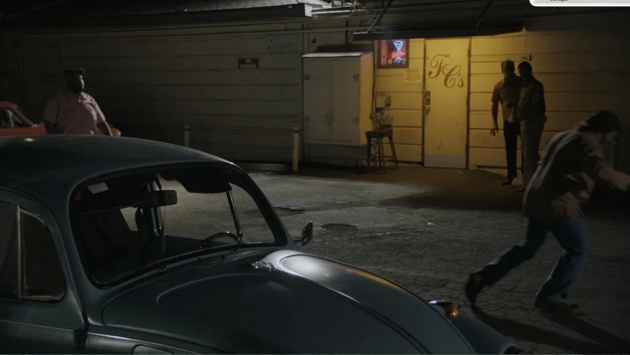 Don't Worry, He Won't Get Far on Foot (2018) movie still: wide — A nighttime parking lot outside a low building with a weathered white facade. A…