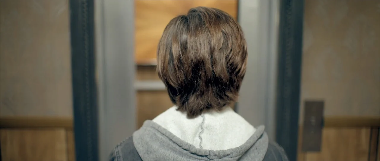 Elevator Game (2023) movie still: medium — An elevator interior with neutral-toned wood paneling on the side walls. A person stands facing away…