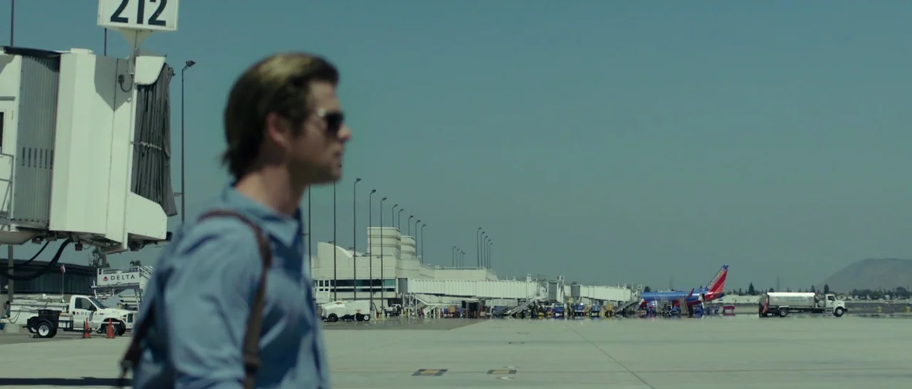 Blackhat (2015) movie still: medium — An airport tarmac stretches out toward distant service buildings and a parked commercial airplane. A man walks…