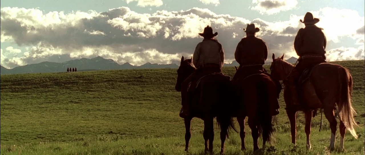 Open Range (2003) movie still: wide — A vast green landscape with rolling hills and a mountain range in the distance. Three cowboys on horseback are…