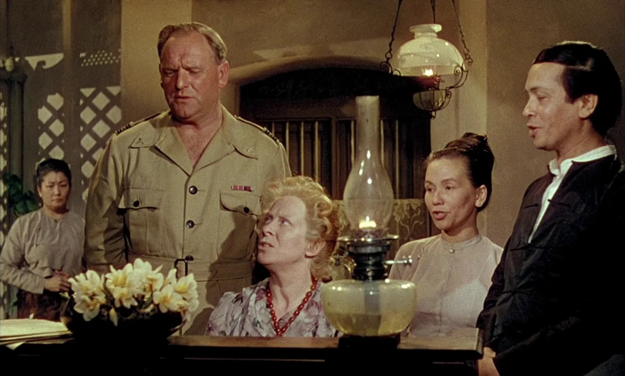 The Purple Plain (1954) movie still: medium — Four individuals gather in a dim room illuminated by a prominent glass oil lamp. The group stands…