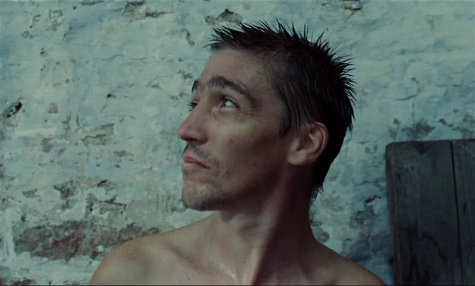 Hadewijch (2009) movie still: closeup — A man stands before a weathered stone wall with chipped light blue paint. The shirtless subject gazes…