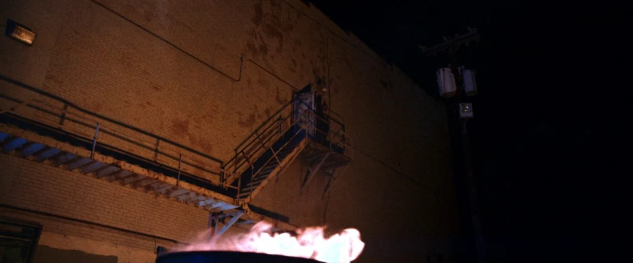 VFW movie still: wide — A dark brick building features a metal fire escape staircase leading to an open door. A metal trash barrel containing an…