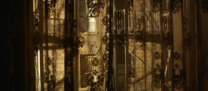 The House of Mirth (2000) movie still: establishing — An urban brick facade with a metal fire escape is viewed through dark lace curtains. The…