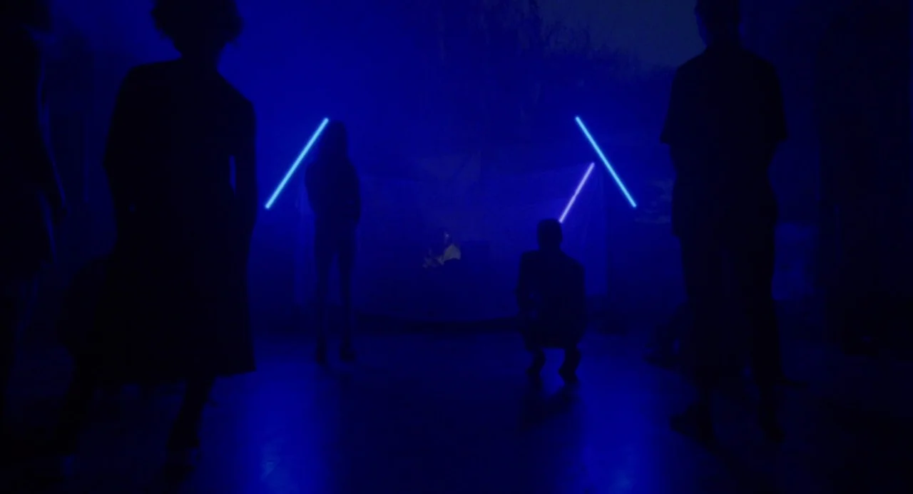 So Pretty (2019) movie still: wide — A dark, blue-lit nightclub room contains several silhouetted figures arranged in front of equipment. One figure…