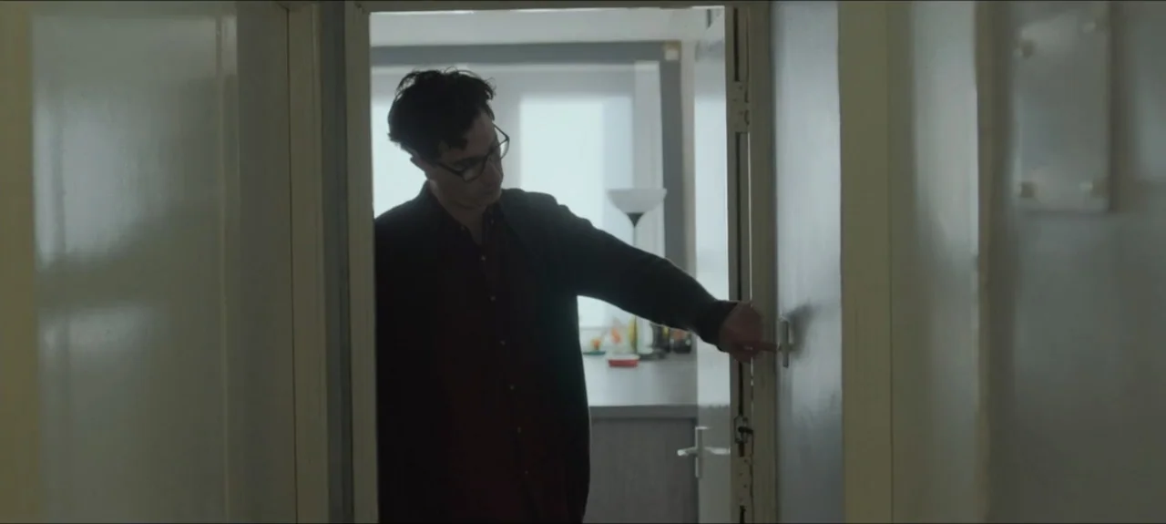 Containment (2015) movie still: medium — A man stands in a door frame connecting a hallway to an adjacent living area. The subject extends his arm…