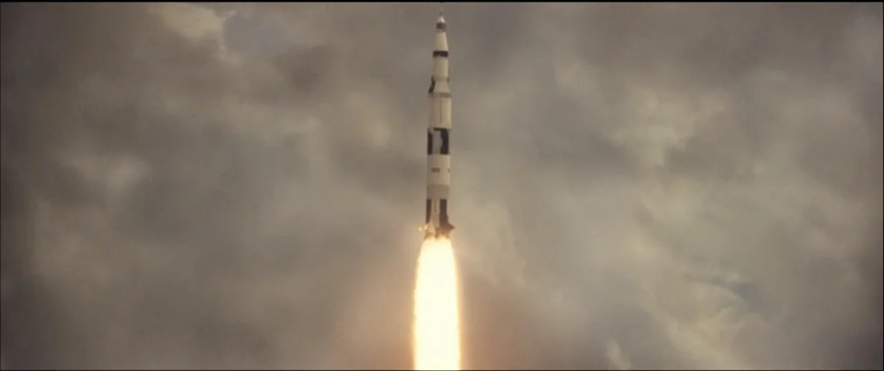 First Man (2018) movie still: wide — A tall Saturn V rocket ascends vertically into a dark, overcast sky filled with clouds. Bright orange flames…