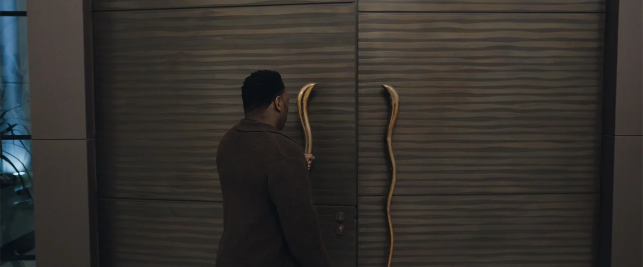 Play Dirty (2025) movie still: medium — A modern interior hallway features a wall clad in dark, horizontal-grained wooden panels. A man in a brown…