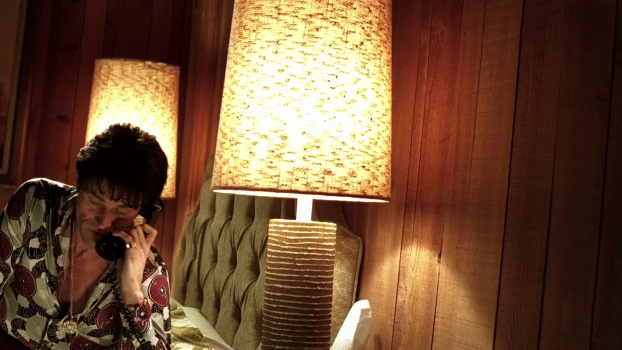 Auto Focus movie still: medium — A 1970s room with vertical wood paneling and a tufted olive fabric headboard. A middle-aged man sits on the bed…