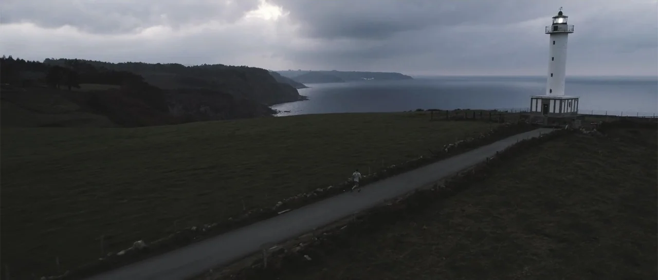 Taking Back the Waves (2017) movie still: wide — A dark, overcast coastal landscape features a tall white lighthouse near the sea cliffs. A lone…