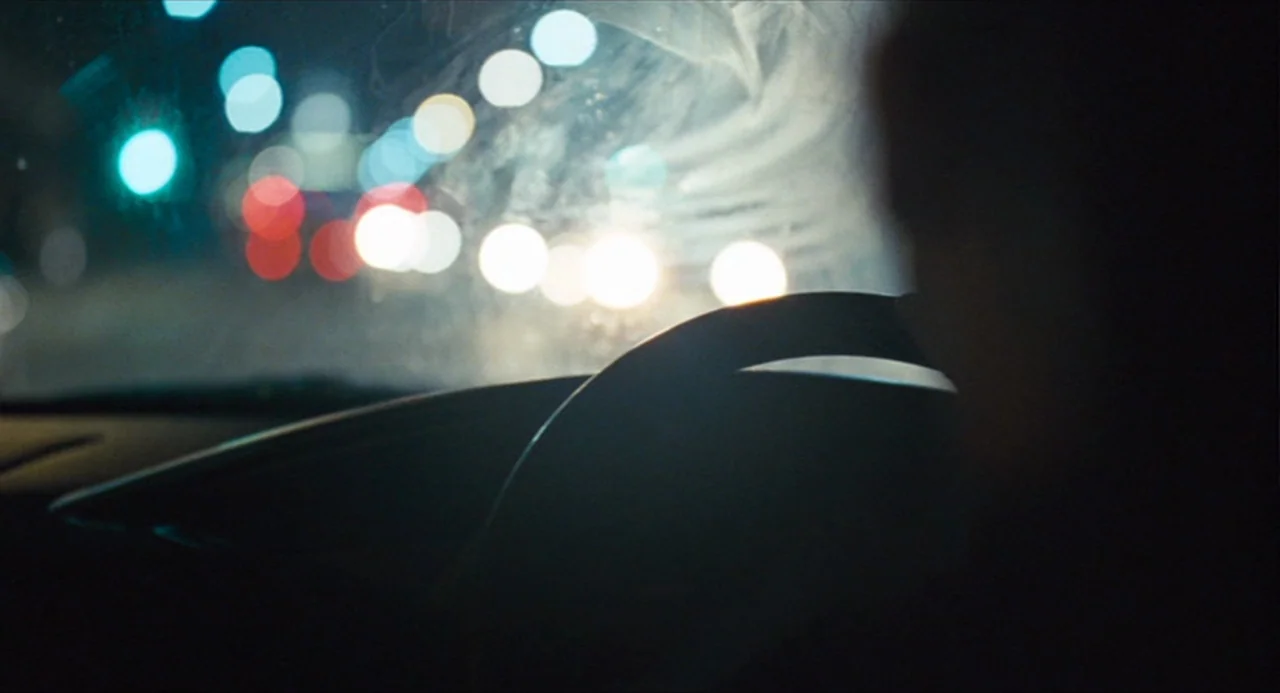 The Angel’s Share (2012) movie still: closeup — Inside a vehicle at night with a blurred view of city lights ahead. A driver is partially visible in…