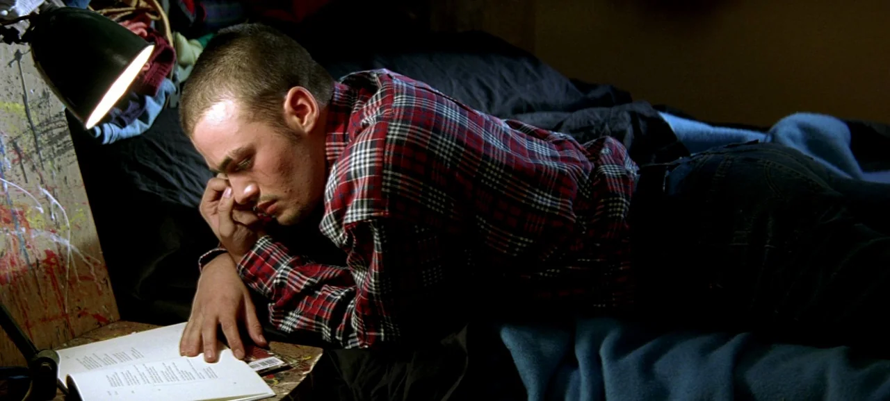 Live Flesh (1997) movie still: medium — A cluttered room features a dark bed with blue blankets and paint-splattered workspace surfaces. A young man…