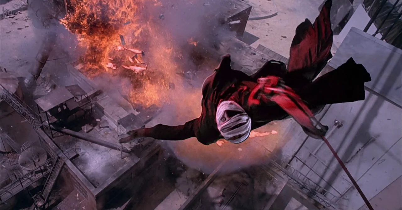 Darkman (1990) movie still: fullbody — An industrial rooftop structure is engulfed in flames and smoke from a recent blast. A man wrapped in…