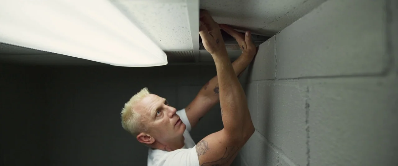 Logan Lucky (2017) movie still: medium — A small room features grey concrete block walls and a drop ceiling. A man reaches up to move a ceiling tile…