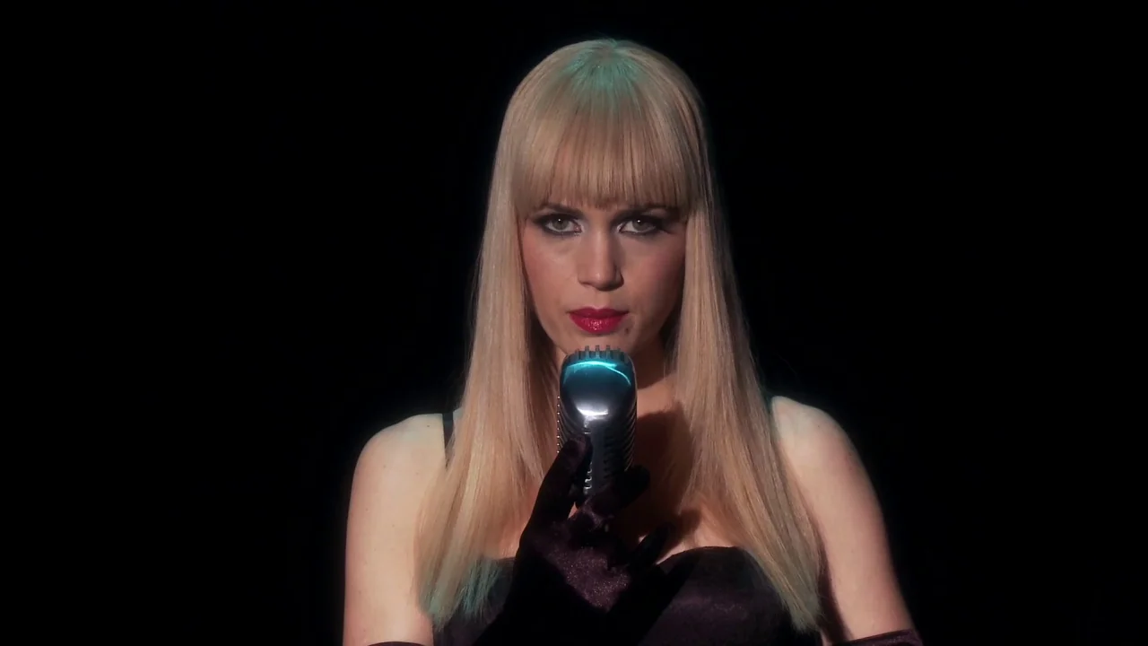 Electra Luxx (2010) movie still: medium — A woman stands before a black backdrop in a dimly lit performance space. She grips a chrome vintage…