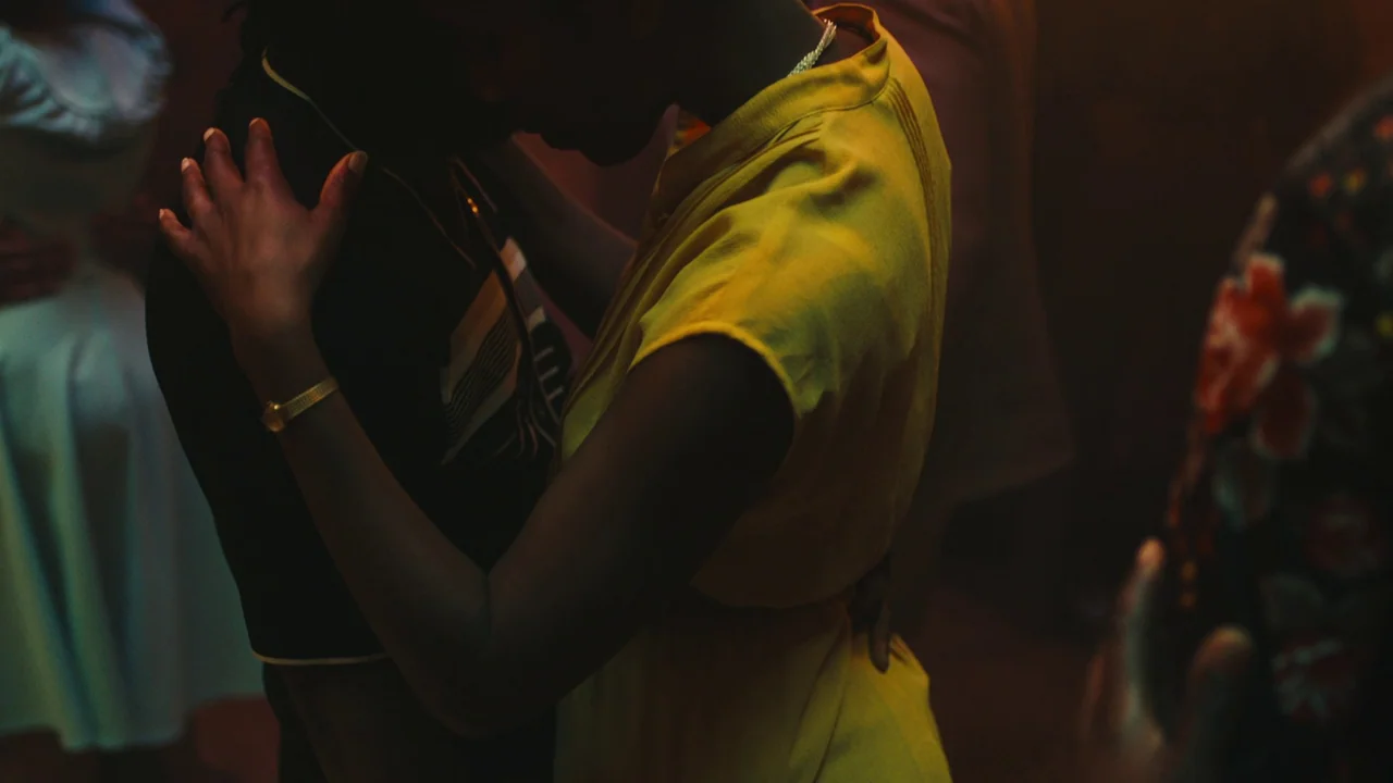 Lover’s Rock (2020) movie still: medium — A dark, dimly lit dance floor filled with people moving in close proximity. A man and a woman in a yellow…