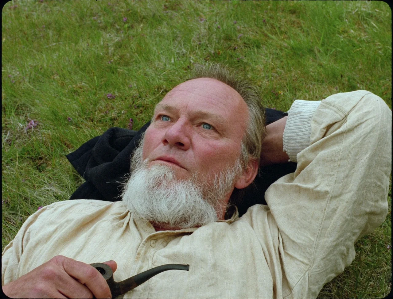 Godland (2022) movie still: closeup — An older man with a white beard rests in a patch of green grass. He holds a dark wooden pipe in his hand while…