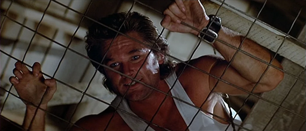 Big Trouble in Little China (1986) movie still: closeup — A man looks upward through a metal wire grate in a dark, confined space. The man grips the…