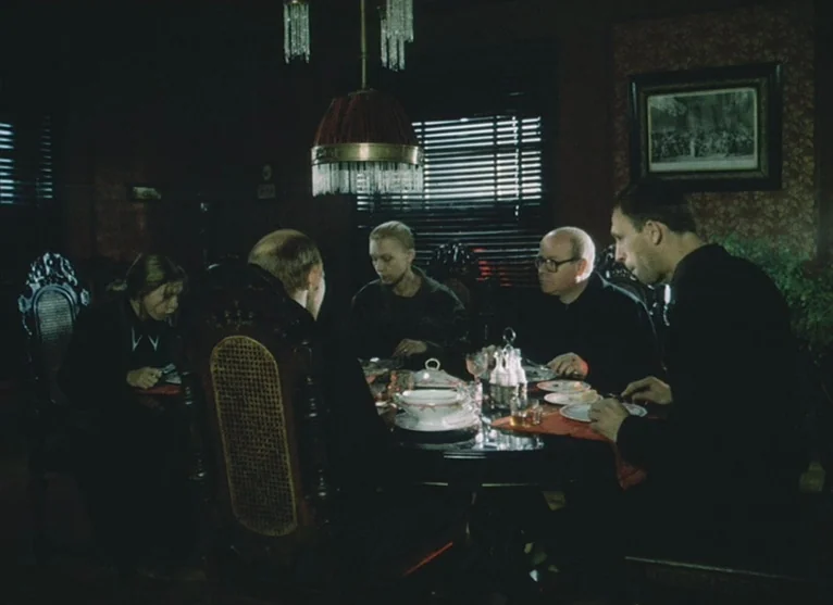 A Visitor to a Museum (1989) movie still: medium — A dimly lit room with dark floral wallpaper and wooden furniture. Four people eat silently around…