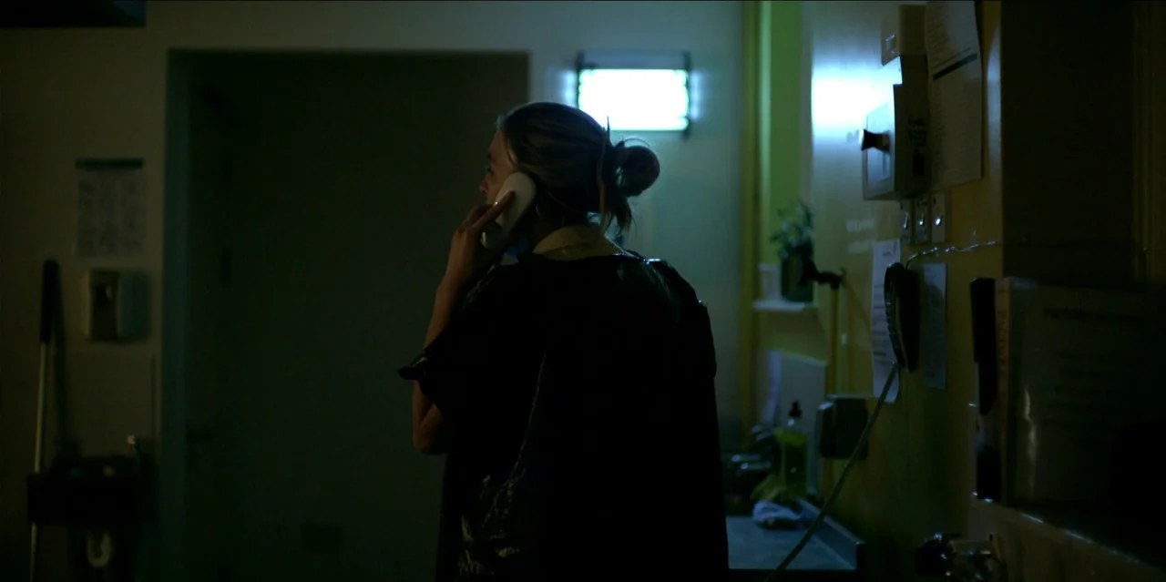 Help (2021) movie still: medium — A dimly lit room features a wall with posted papers and a glowing light fixture. A woman standing in silhouette…