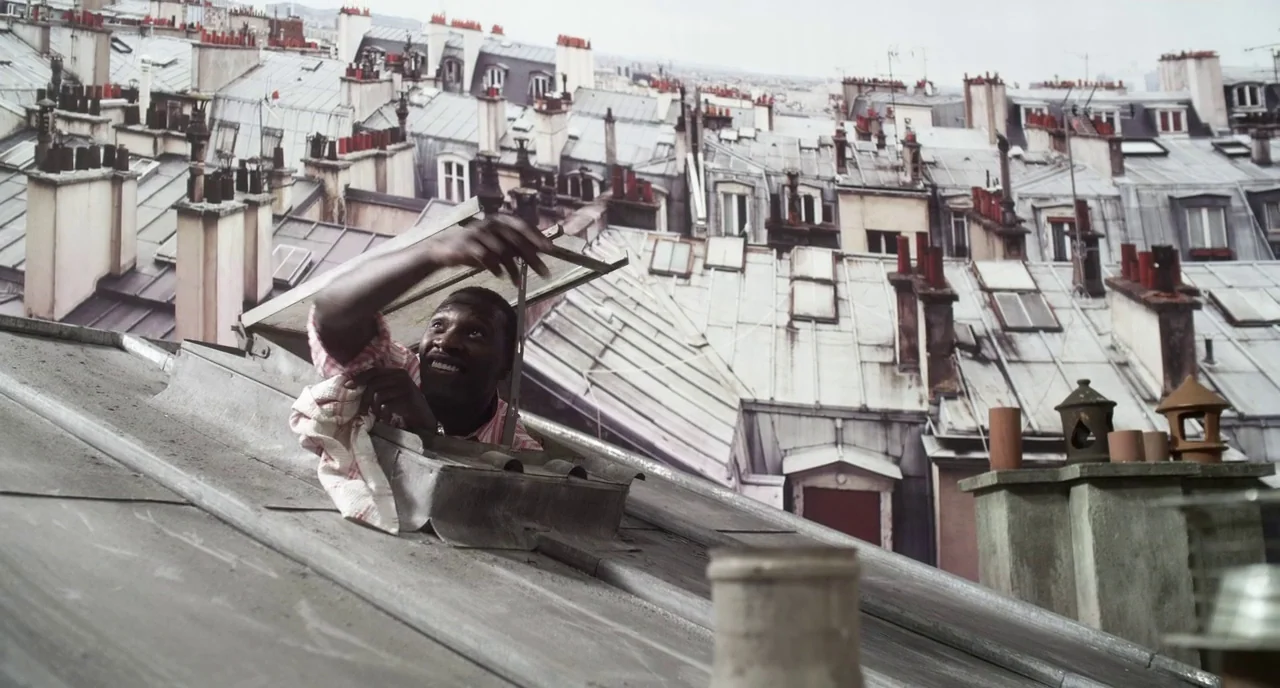 Mood Indigo (2013) movie still: medium — A dense urban rooftop setting with multiple chimney stacks and grey metallic roof panels. A man emerges…