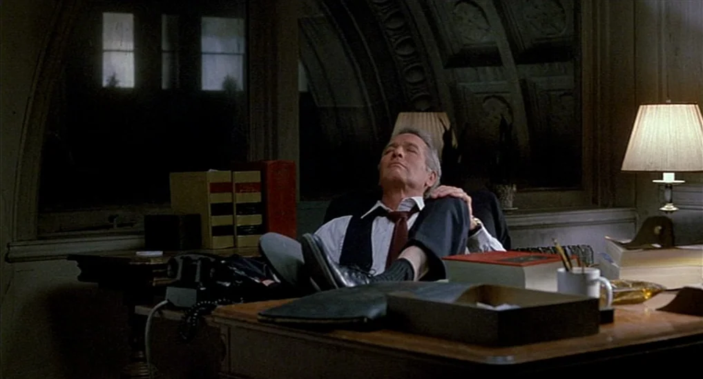 The Verdict (1982) movie still: medium — A dimly lit room contains a cluttered wooden desk with books, a phone, and lamp. The man rests with his…