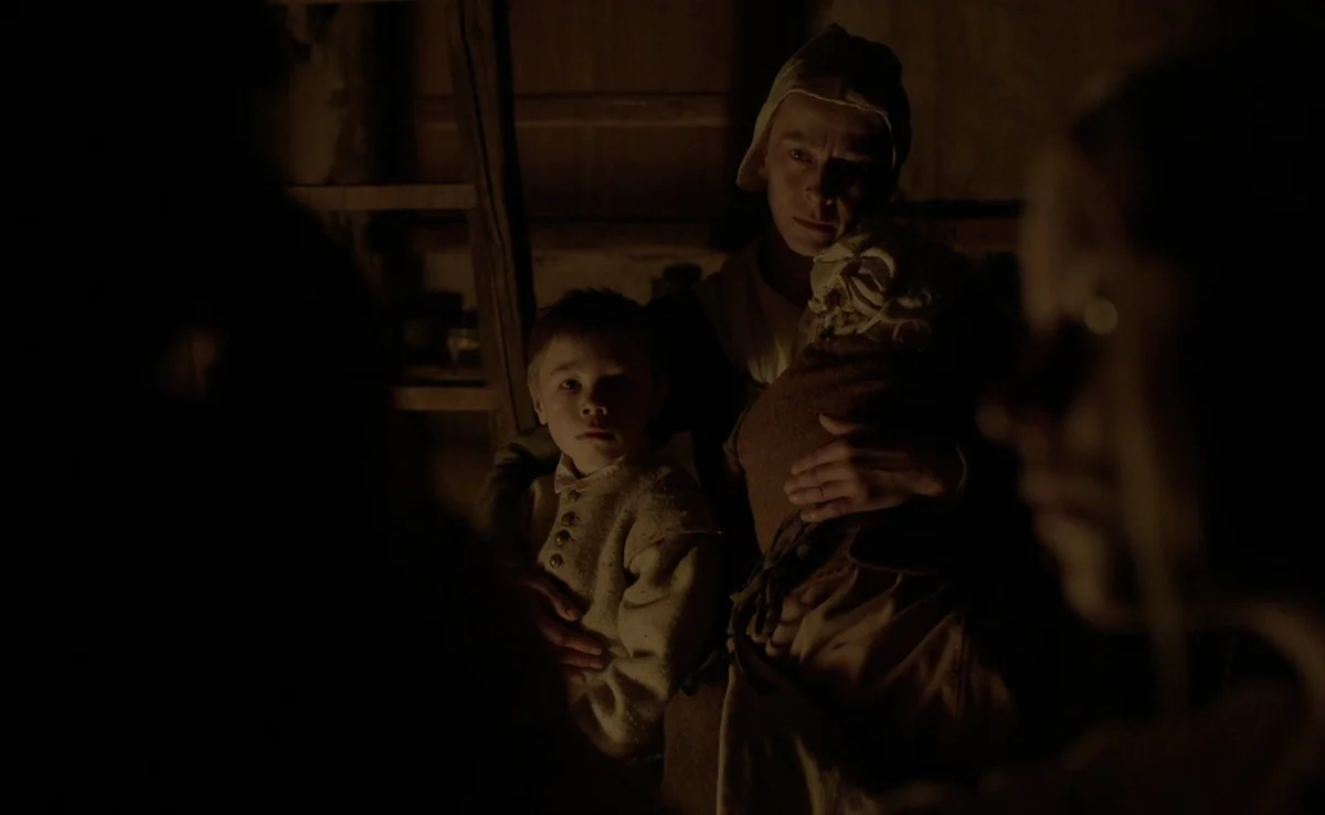 The Witch (2015) movie still: medium — A dark, cramped wooden cabin features a ladder leaning against a rough-hewn wall. A woman holding two young…