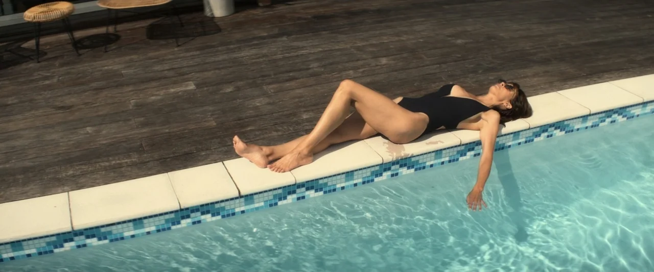 I Love America (2022) movie still: fullbody — A wooden deck sits beside a blue tiled swimming pool in bright daylight. A woman in a black swimsuit…