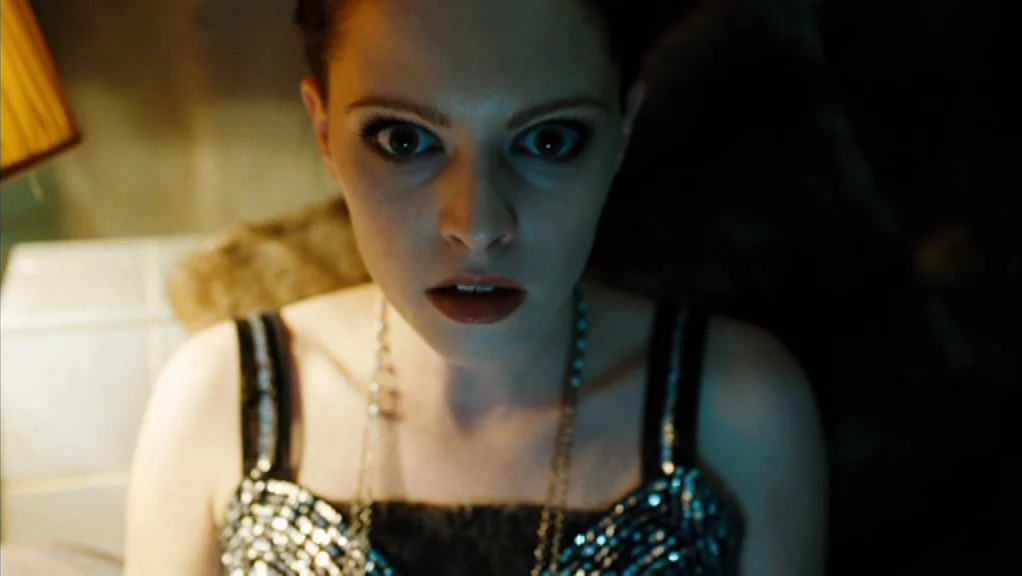 We Are The Night (2010) movie still: closeup — A dimly lit room with dark shadows surrounding a woman in a sequined top. The woman gazes ahead with…