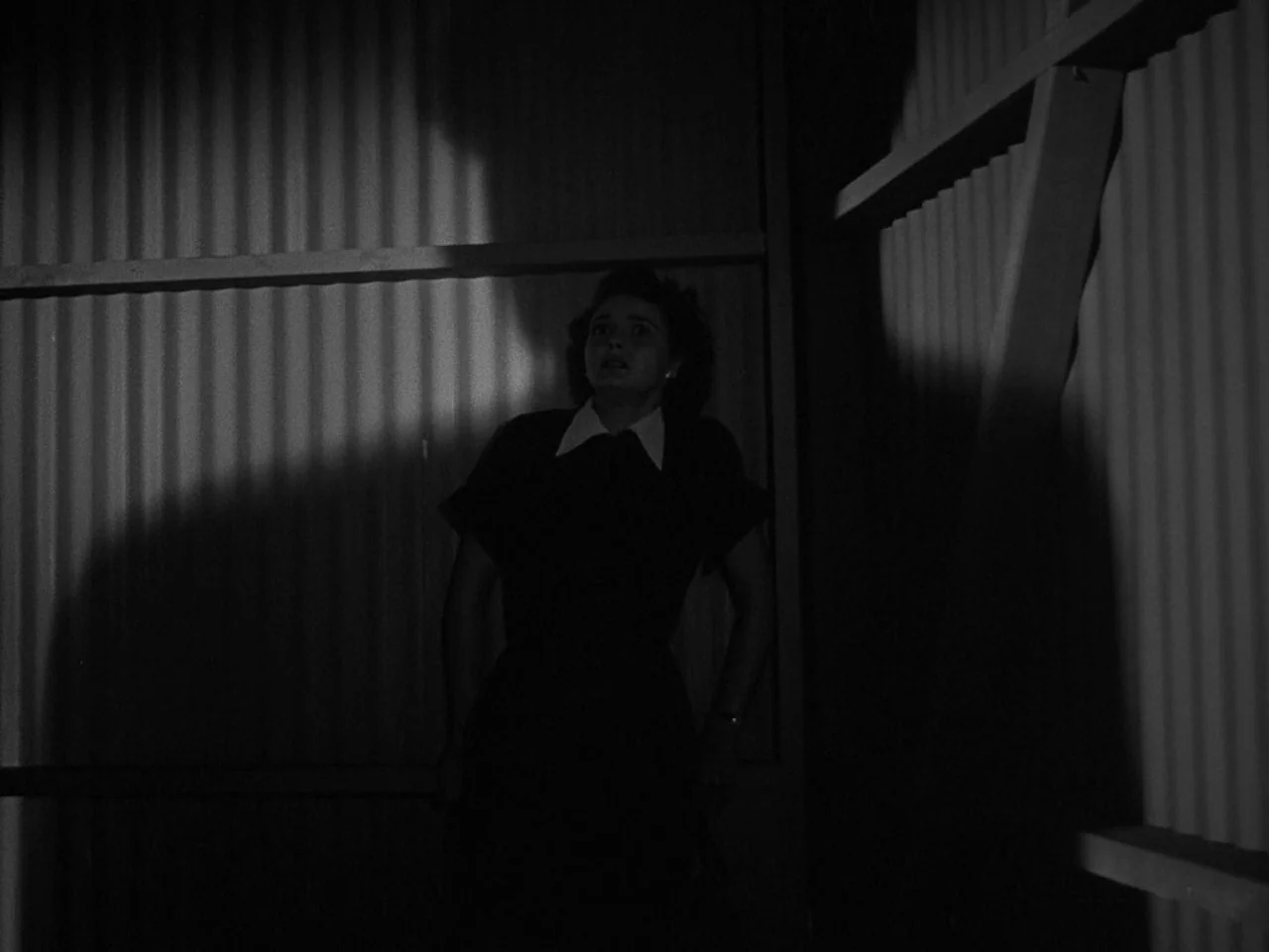 The Day the Earth Stood Still (1951) movie still: medium — A woman stands in a dark corner with corrugated metal walls. She looks upward with a…