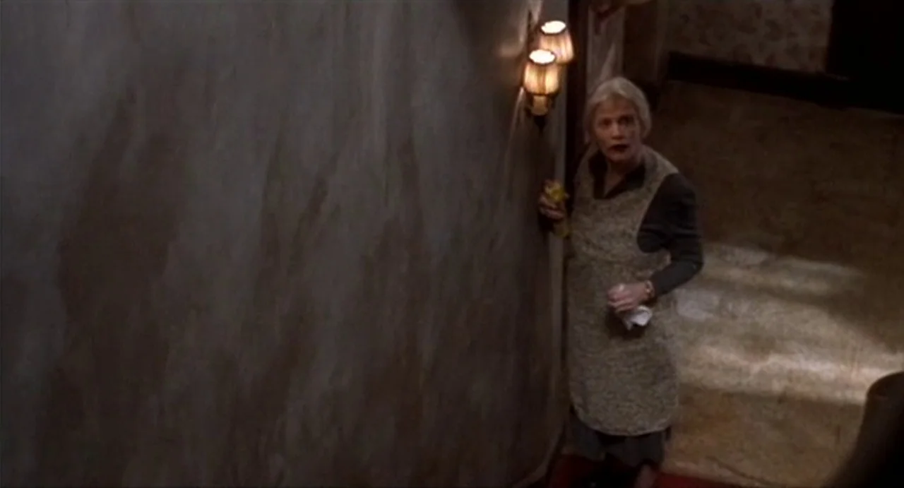 The Annihilation of Fish (1999) movie still: medium — A dim interior staircase landing features plain walls and a wall-mounted brass light fixture.…
