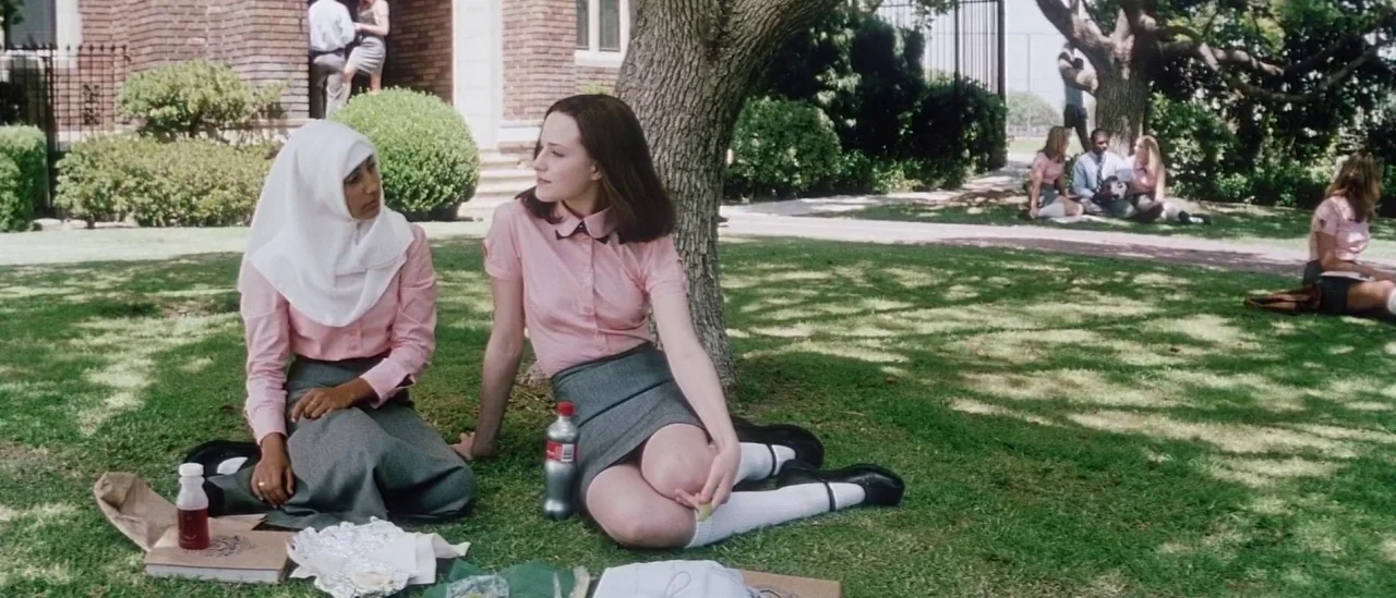 Pretty Persuasion (2005) movie still: fullbody — Two teenage girls sit on a manicured green lawn near a brick building. They are positioned under…