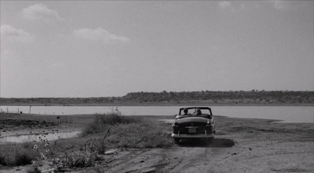 The Last Picture Show (1971) movie still: wide — A wide, sparse landscape features a dirt road leading towards a calm lake. A vintage convertible…