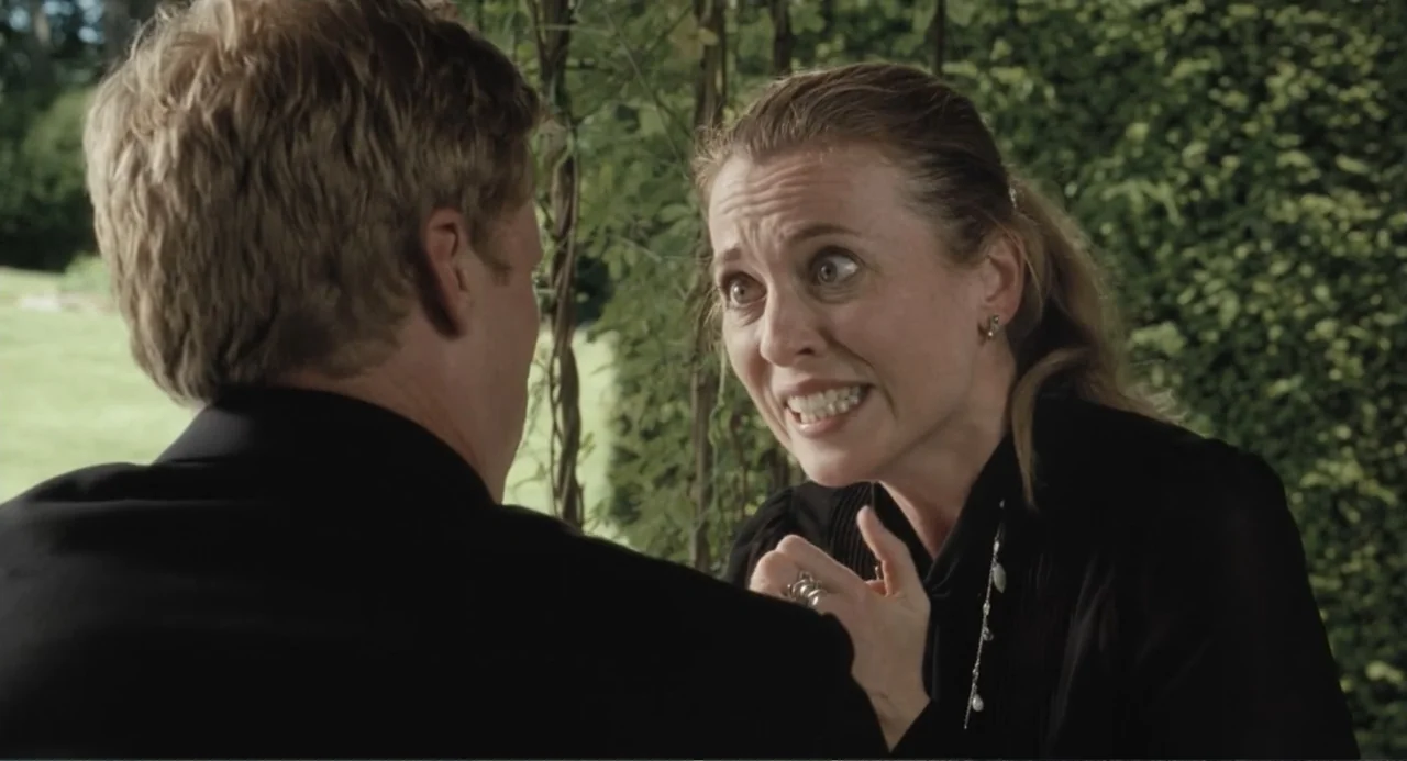 Death at a Funeral (2010) movie still: medium — A green outdoor space with dense hedges behind a seated couple at a table. A woman leans over the…