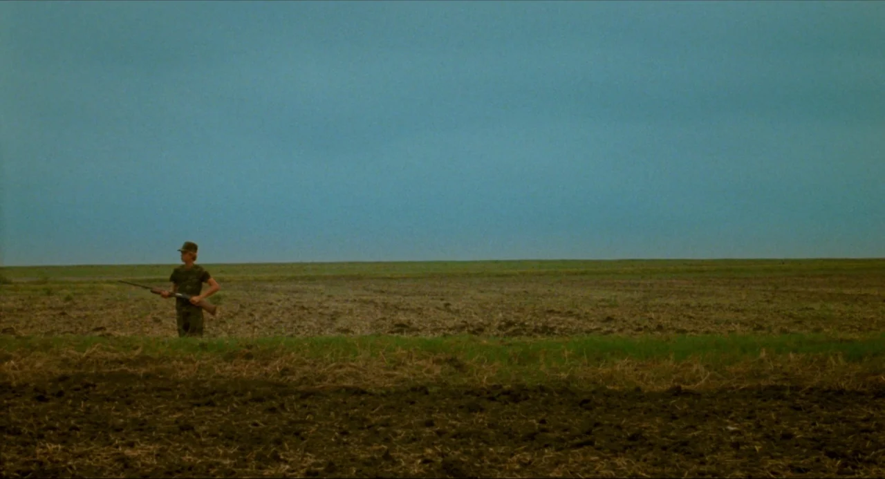 True Stories (1986) movie still: wide — A flat, expansive agricultural field stretches to the horizon under a pale sky. A lone person in camouflage…