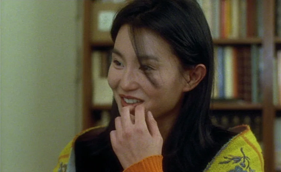 Irma Vep (1996) movie still: closeup — An interior space features a wooden bookshelf filled with books in the blurred background. A young woman with…